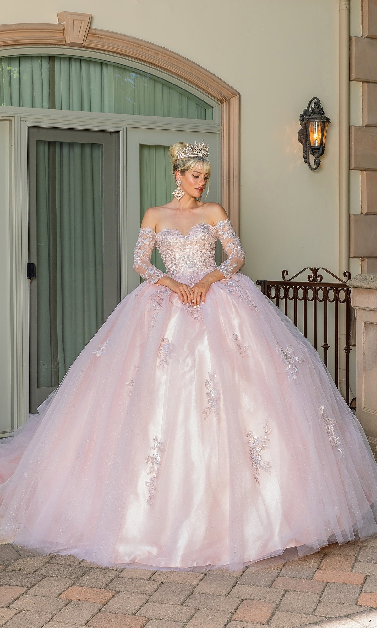 A woman in the Blush Pink Quinceanera Dress 1674 with Sleeves and a crown stands by a building’s large window and balcony, her elegant look highlighted by the gown’s ornate design and soft blush pink color.